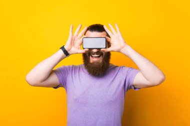 Sarı duvar arkasında duran yakışıklı, sakallı, duygusal adamın fotoğrafı. Telefonla gözleri kapatıyor..