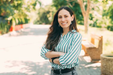 Kollarını kavuşturup parkta poz veren güzel bir kız. Dalgalı saçlı, açık havada duran ve kameraya bakan genç ve güzel bir kadın..