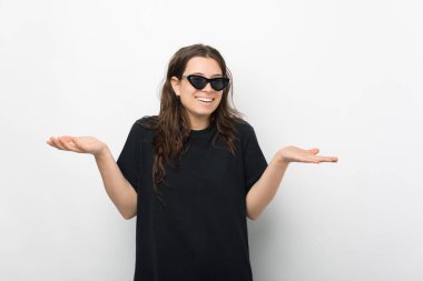 Siyah tişört giyen, güneş gözlüklü, kolları ve elleri havada, kafası karışmış genç bir kadın.