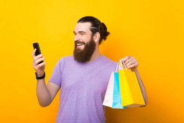 Alışveriş torbalarıyla yakışıklı sakallı adam cep telefonu kullanıyor ve sarı arka planda gülümsüyor.