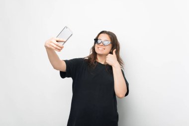 Genç bir kadının güneş gözlüklü selfie 'sini çekerken başparmağını kaldırıp gülümsediği güzel bir fotoğraf. .