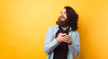 Yakışıklı genç sakallı hippi akıllı telefon kullanıyor ve sarı arka planda gülümsüyor..