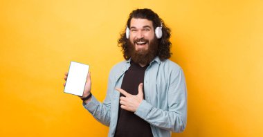 Beyaz kablosuz kulaklık takan ve tabletteki boş ekrana bakan neşeli sakallı hippi..