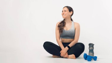 Fit genç bir kadın kelebek şeklinde oturuyor, halterlerin yanında poz veriyor ve beyaz arka planda bir su şişesi var..