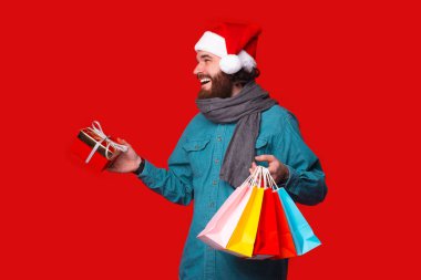 Noel Baba şapkalı sakallı adam, alışveriş torbaları ve arkadaşlar için hediyeler, kırmızı arka plan.