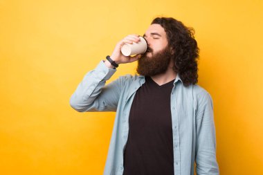 Sıradan giyinmiş, sarı arka planda sabah kahvesini içen sakallı adamın portresi.