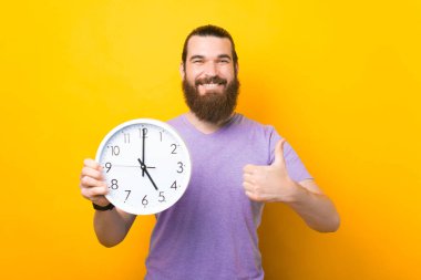Mutlu sakallı adam elinde yuvarlak bir saat tutuyor ve sarı arka planda baş parmağını gösteriyor..