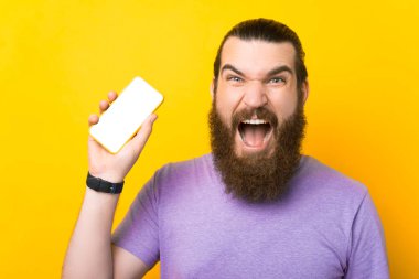 Genç sakallı adam telefonunu tutuyor ve sarı arka planda bir stüdyoda kameraya bağırıyor..