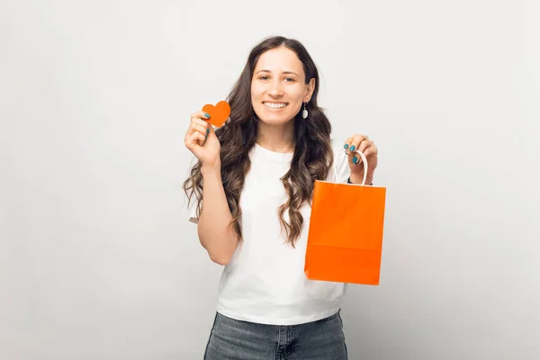 Uśmiechnięta kobieta trzyma pomarańczowy worek na zakupy i serce na białym tle. — Zdjęcie stockowe