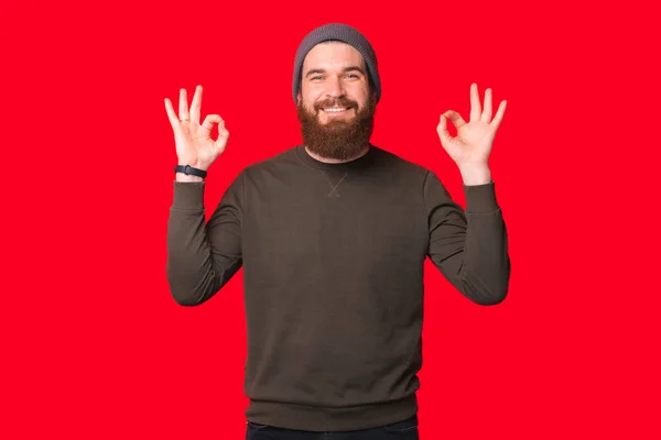 Boldog szakállas férfi mutatja rendben gesztus felett piros háttér. — Stock Fotó
