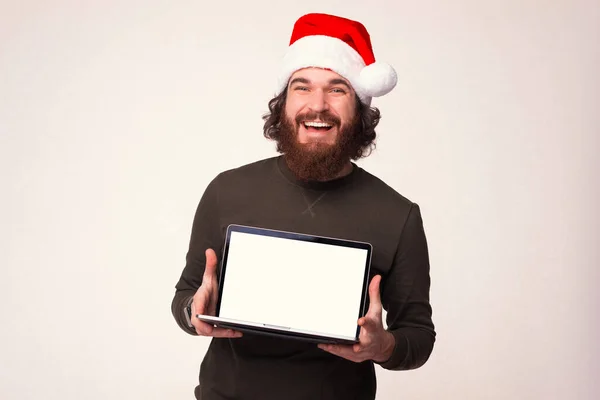 クリスマスの帽子をかぶった若い髭の男がノートパソコンを持っていて、カメラにスクリーンを見せている。. — ストック写真