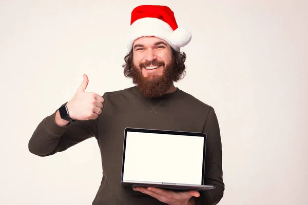 Χαμογελώντας άνθρωπος φορώντας χριστουγεννιάτικο καπέλο δείχνει τον αντίχειρα και κρατώντας το laptop του με την οθόνη που παρουσιάζονται. — Φωτογραφία Αρχείου