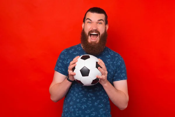 Gritando homem barbudo alegre está segurando uma bola de futebol com as duas mãos. — Fotografia de Stock
