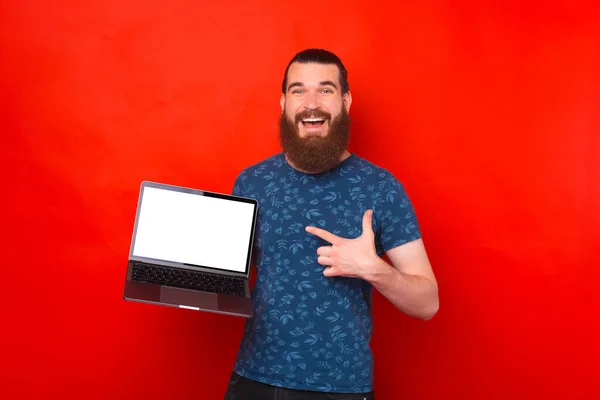 Glücklicher bärtiger Mann zeigt auf den weißen leeren Bildschirm vor rotem Hintergrund. — Stockfoto