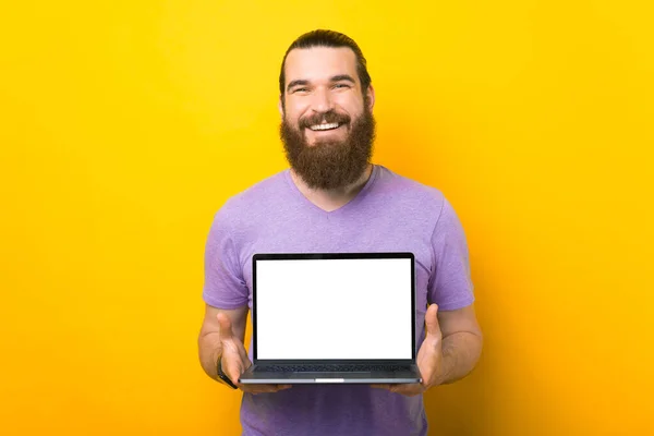 Maravilloso hombre barbudo está sosteniendo una computadora portátil de pantalla en blanco a la cámara. — Foto de Stock