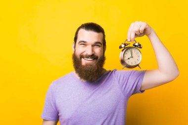 Gülümseyen sakallı adam sarı arka planda çalar saati tutuyor..