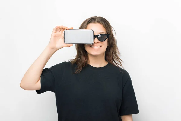 Glimlachende vrouw bedekt een oog met haar telefoon, terwijl staan over witte achtergrond. — Stockfoto