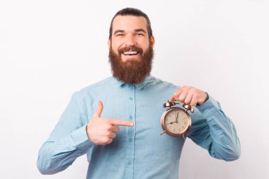 Kameraya gülümseyen sakallı adam çalar saati işaret ediyor..