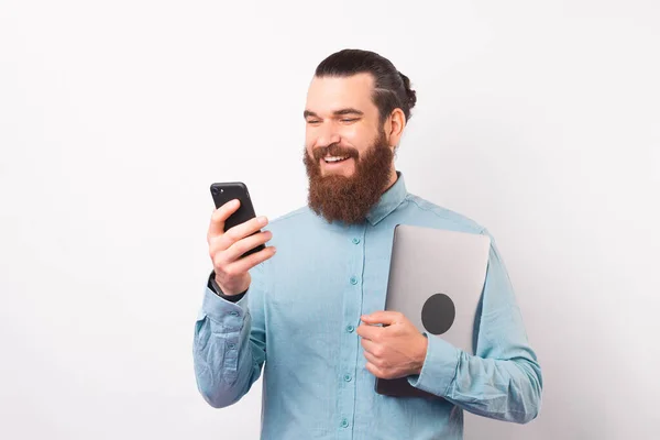 Sourire homme barbu utilise son téléphone tout en tenant ordinateur portable. — Photo