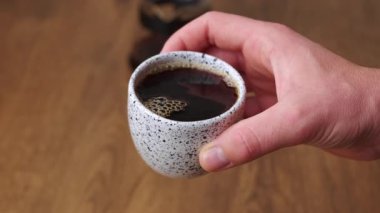 Bir el, güzel benekli seramik bir fincanı nazikçe tutar. Kırsal ahşap bir yüzey üzerinde taze demlenmiş kahveyle dolu.
