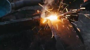 parlak kaynak yayı çeliği kesiyor, meşalenin yakın çekimi mavi-beyaz işaret fişeği ile eriyen kalın plaka, uçan kıvılcımlar ve kor duşu, dumanlı atölye