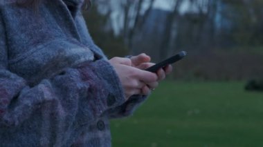 Beyaz kadının elleri akıllı telefonu yakından çekiyor, yün kol görünür, yeşil çimenlik arka plan, başparmak kaydırma.,