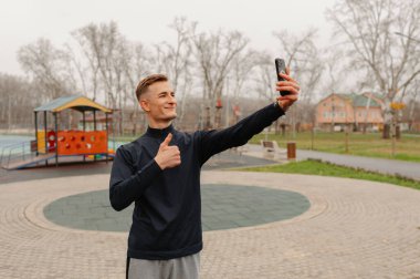 Kafkasyalı bir adam çocuk parkında selfie çekiyor. Kapüşonlu, atletik giysili ve cep telefonlu olarak başparmak kaldırıyor.