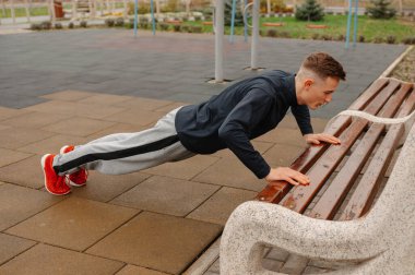 Kafkasyalı adam bankta eğik şınav çekiyor, sabit çekirdek odaklı set, gri eşofman ve kırmızı ayakkabılar, açık havada