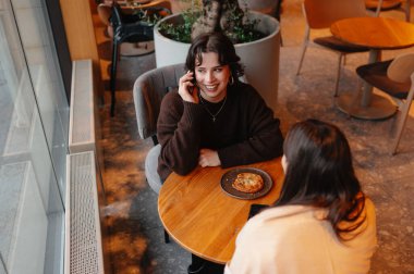 Kahvede gülümseyen genç bir kadın hamur işini ahşap masada paylaşıyor, pencereden gelen sıcak gün ışığı, saksı bitkisi