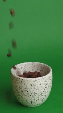 Fasulyelerle dolu seramik kupa. Kavrulmuş kahve çekirdekleri canlı yeşil zemine karşı benekli fincana dökülüyor.,