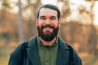 Sonbahar parkında gülümseyen sakallı adam, sırt çantalı gezgin altın saat günışığının ve yumuşak Bokeh 'in tadını çıkarıyor.,