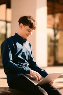 Genç beyaz öğrenci kampüste dizüstü bilgisayar kullanıyor. Sunlit Avlu, odaklanmış ifade, donanma gömleği, daktilo.