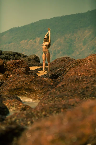 Молодая девушка в бикини с красивым телом и солнцезащитными очками возле красных камней на пляже — стоковое фото