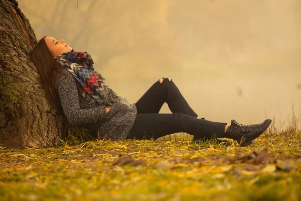 Einsame Frau, die sich an einem nebligen Herbsttag unter dem Baum am Wasser ausruht. einsame Frau genießt die Landschaft im Herbst. Herbsttag. Mädchen sitzt auf Gras horizontale Bild. — Stockfoto