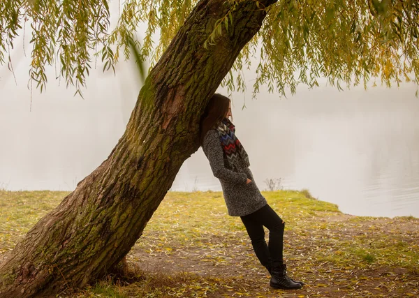 Einsame Frau, die sich an einem nebligen Herbsttag unter dem Baum am Wasser ausruht. einsame Frau genießt die Landschaft im Herbst. Herbsttag. Mädchen sitzt auf Gras horizontale Bild. — Stockfoto