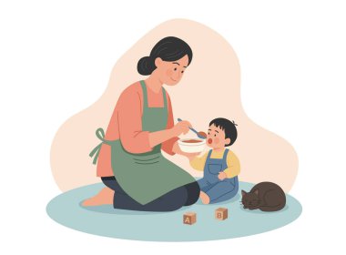 Yeşil önlüklü bir annenin diz çöküp küçük çocuğunu kaşıkla beslediğinin sıcak bir örneği. Uyuyan kedi ve tahta alfabe blokları rahat bir ev sahnesi yaratır..