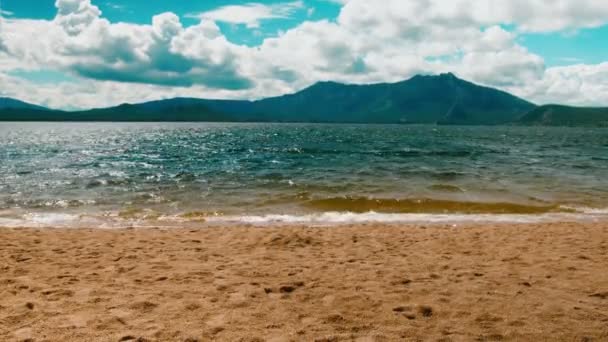 Onde che si infrangono dolcemente sulla tranquilla spiaggia sabbiosa. sfondo di montagna. Filmati 4K — Video Stock