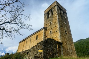 Bir saati ve kemerli girişi olan uzun taş çan kulesi, yeşilliklerle çevrili ve parlak mavi gökyüzüne karşı kurulmuş..