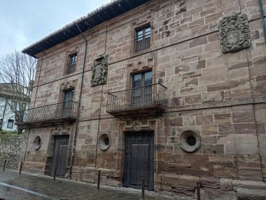 Köyde soylu armaları, balkonları ve dairesel pencereleri olan büyük taş bir ev.