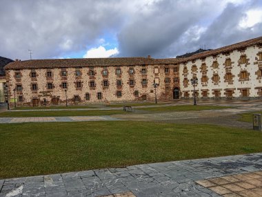 Eşit aralıklı pencere ve bayrakları olan büyük dikdörtgen bina, desenli bir taş ve çimen plazasının arkasına kurulmuş.