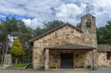 Bulutlu gökyüzünün altında yeşil çam ağaçları arasında çan kulesi olan tarihi taş şapeli.