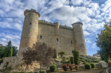 İkonik Castillo de Torija, sağlam taş duvarlar ve silindirik kuleler, canlı mavi bir gökyüzüne karşı dimdik ayakta duruyor ve yeşilliklerle çevrili..