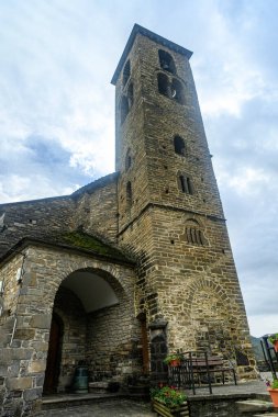 Uzun bir çan kulesi ve kırsal detaylar ve bulutlu gökyüzü ile çevrili kavisli bir girişi olan Romanesk taştan bir kilise..