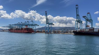 Mavi gökyüzünün altında feribot, kargo gemisi, vinç ve depolama tankları olan Algeciras Limanı.