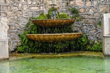 Yeşil bitkilerle çevrili, oyulmuş yüzlü ve çağlayan sularla süslü bir çeşme.
