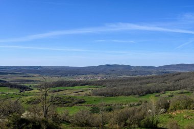 Dağların panoramik manzarası, yeşil tarlalar, ve parlak mavi gökyüzünün altındaki uzak karlı tepeler..