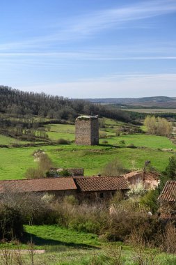 Yeşil tepeler ve geleneksel çatılarla çevrili kırsal bir alanda eski bir savunma taşı kulesi..