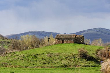 Çıplak ağaçlar ve fırtınalı bulutlarla kaplı bir tepedeki antik taş şapel.