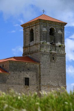 Bulutlu bir gökyüzünün altında, yüksek yeşil çimlerin arkasında yükselen kırmızı çatılı Taştan köy kilisesi.