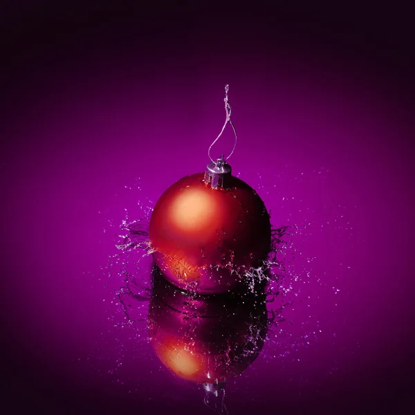 Red Christmas bauble on purple background with splashes of water — Stock Photo, Image